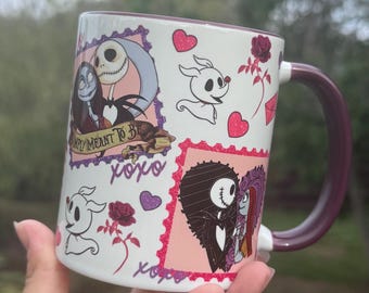 Op de afbeelding: Witte keramische mok met een bordeauxrode handgreep en rand, met personages uit The Nightmare Before Christmas. Het ontwerp bevat Jack Skellington, Sally, Zero, harten en de tekst "Simply Meant to Be" en "xoxo".