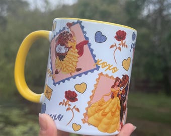 Op de afbeelding: Witte keramische mok met een gele handgreep en binnenrand. De mok toont illustraties van een stel in gele en rode kleding, omringd door harten, rozen en de tekst "xoxo".