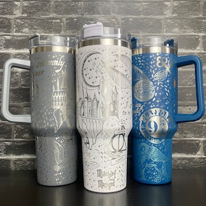 May include: Three stainless steel travel mugs with handles, featuring engraved designs. One is grey with a castle, one is white with a moon and stars, and one is blue with a winged key and Platform 9 3/4. Each mug has a clear lid.