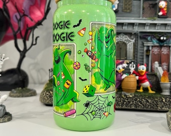 Op de afbeelding: Een lichtgroene glitterglazen beker met een bamboe deksel, voorzien van een grafische print van een groen personage, snoep, dobbelstenen en spinnenwebben. De tekst "OOGIE BOOGIE" staat op de zijkant van de cilindervormige beker met platte bodem.