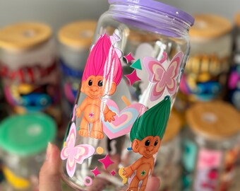 Op de afbeelding: Transparante glazen pot met een paarse deksel, versierd met cartoon trollen, vlinders, harten en sterren. De pot heeft een brede opening en wordt in een hand gehouden. Andere potten zijn zichtbaar op de achtergrond.