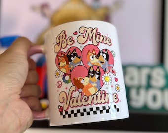 Op de afbeelding: Een witte keramische mok met een roze handvat. De mok heeft een retro-stijl grafische afbeelding met de tekst "Be Mine Valentine" en cartoonhonden in harten.