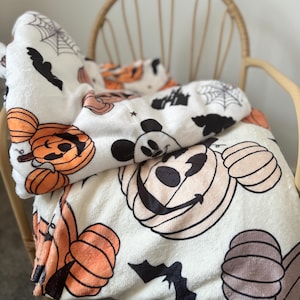Op de afbeelding: Witte fleece deken met een Halloween-thema met Mickey Mouse en pompoenen. De deken heeft een herhalend patroon van Mickey Mouse's gezicht met een pompoen als hoofd, pompoenen, vleermuizen en spinnenwebben.