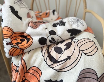 May include: White fleece blanket with a Halloween theme featuring Mickey Mouse and pumpkins. The blanket has a repeating pattern of Mickey Mouse's face with a pumpkin for a head, pumpkins, bats, and spiderwebs.