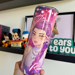 Op de afbeelding: Een roestvrijstalen beker met een anime-stijl illustratie van een vrouw met paars haar en een witte trui. De beker heeft de tekst "Joey Rumi" en een neonroze "Rumi" aan de zijkant. De achtergrond is een mix van roze, paars en blauw.