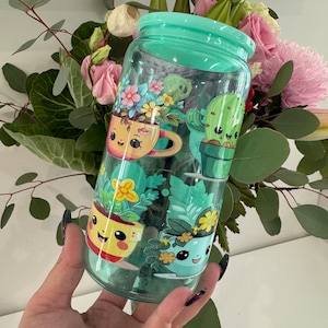 Op de afbeelding: Een doorzichtige herbruikbare waterfles van plastic met een groene dop en een schattig cartoonontwerp met potplanten met gezichten. De planten zijn een cactus, een succulent en twee verschillende soorten bloemen.