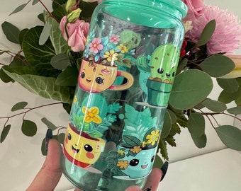 Op de afbeelding: Een doorzichtige herbruikbare waterfles van plastic met een groene dop en een schattig cartoonontwerp met potplanten met gezichten. De planten zijn een cactus, een succulent en twee verschillende soorten bloemen.