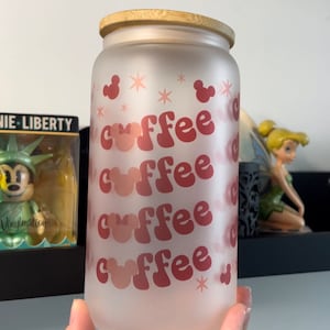 Op de afbeelding: Matglazen beker met een bamboe deksel. De beker heeft het woord "coffee" herhaald in een retro lettertype, met Mickey Mouse-hoofdvormen verwerkt in het ontwerp. Roze sterren en Mickey Mouse-vormen versieren het glas.