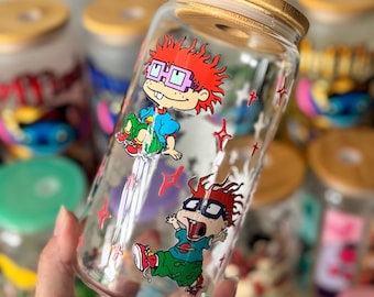 Op de afbeelding: Een heldere glazen beker met een bamboe deksel, met cartoonafbeeldingen van een personage met rood haar en een bril. De beker heeft een cilindrische vorm en is versierd met rode sterren.