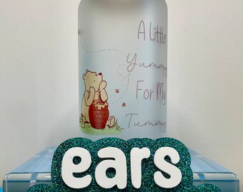 Op de afbeelding: Een mat glazen pot met een houten deksel heeft een afbeelding van Winnie de Poeh en de tekst "A little yummy for my tummy". De pot staat op een blauw en groen glitteroppervlak met de tekst "ears to you" in wit.