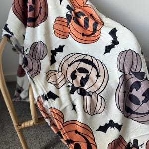 Op de afbeelding: Witte fleece deken met een Halloween-thema print met oranje, bruine en zwarte pompoenen met Mickey Mouse oren en vleermuizen.