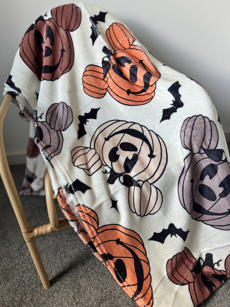 Op de afbeelding: Witte fleece deken met een Halloween-thema print met oranje, bruine en zwarte pompoenen met Mickey Mouse oren en vleermuizen.