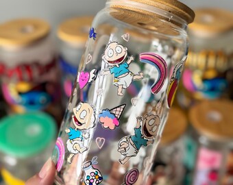 Op de afbeelding: Transparante glazen pot met een bamboe deksel, versierd met tekenfilmfiguren, regenbogen, harten en snoep. De pot is versierd met kleurrijke afbeeldingen van een populaire tekenfilm. De pot wordt in een hand gehouden.