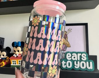 Op de afbeelding: Een doorzichtige glazen beker met een roze deksel en een holografisch zwart-wit schaakbordpatroon. Het ontwerp bevat het woord MAMA dat drie keer wordt herhaald in een golvend roze lettertype, geaccentueerd door kleine lachende bloemiconen.