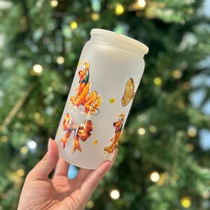 May include: Frosted glass tumbler featuring Pluto and a bow design. The tumbler has a white lid and is decorated with cartoon images of Pluto, a bone, and a bow. The background is a blurred Christmas tree.