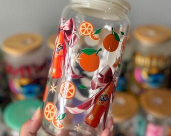 Op de afbeelding: Een heldere glazen pot met een witte deksel, versierd met sinaasappelschijfjes, sinaasappels en Fanta frisdrankflessen. De pot heeft een rood strikdesign en steraccenten. Andere potten zijn zichtbaar op de achtergrond.