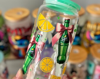 Op de afbeelding: Een doorzichtige glazen beker met een felgroen deksel, versierd met afbeeldingen van groene Sprite-flessen met roze strikken en gele citroenschijfjes, geaccentueerd door kleine groene sterretjes verspreid over het oppervlak van het glas.