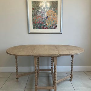 May include: A light brown wooden drop-leaf table with an oval top and four barley twist legs. The table features a central fixed section with two hinged side leaves that can be folded down. The wood grain is visible across the surface.