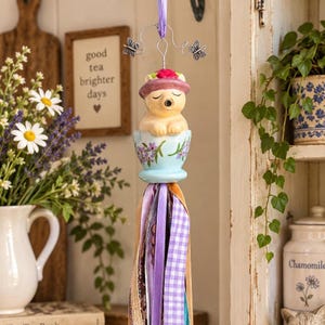 May include: A hanging ornament featuring a ceramic bear wearing a pink hat, seated inside a light blue teacup decorated with purple flowers. The ornament is suspended by a purple ribbon and silver wire with butterfly accents, finished with a long tassel of assorted ribbons in shades of purple, brown, and white.