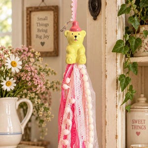 May include: A hanging decoration featuring a yellow bear figurine wearing a pink hat, suspended from a pink ribbon. Below the bear is a long, layered tassel made of various pink and white ribbons, lace, pom-pom trim, and a string of small beads.