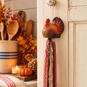 May include: A hanging turkey ornament featuring a painted ceramic turkey figure in shades of brown, orange, and red. Attached to the base of the turkey is a long, decorative tassel made of multiple strands of lace, ribbon, and trim in autumn colors.