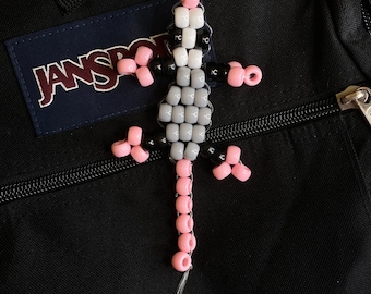 May include: A pink, black, and white beaded lizard keychain hanging on a black Jansport backpack.