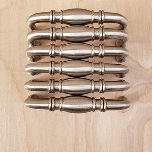 May include: Six silver cabinet pulls with a rounded, slightly curved design. The pulls have a brushed finish and are arranged in a stack.