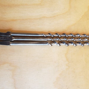 May include: Three silver drill bits with a twist design, laid out on a light brown wooden surface.