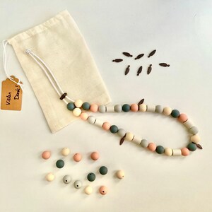May include: A long necklace made of round and cylindrical silicone beads in shades of sage green, dusty pink, cream, and grey, featuring small dark brown leaf-shaped charms. Includes a beige fabric drawstring pouch and a tag reading Vielen Dank.