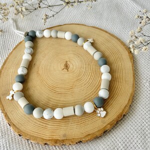 May include: A beaded necklace featuring a mix of round and cylindrical beads in shades of white, gray, and marble patterns. The necklace includes two small metal charms, one shaped like a bone and the other shaped like a paw print, attached to the strand.