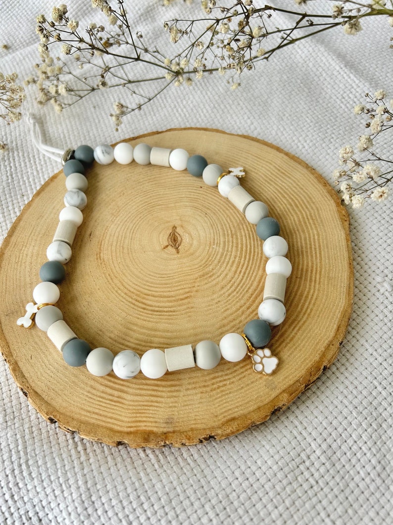 May include: A beaded necklace featuring a mix of round and cylindrical beads in shades of white, gray, and marble patterns. The necklace includes two small metal charms, one shaped like a bone and the other shaped like a paw print, attached to the strand.