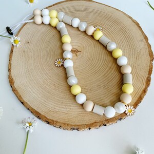 May include: A beaded lanyard featuring a sequence of round and cylindrical beads in shades of white, cream, beige, and yellow. The accessory includes small metal daisy charms with white petals and yellow centers, strung on a white cord.