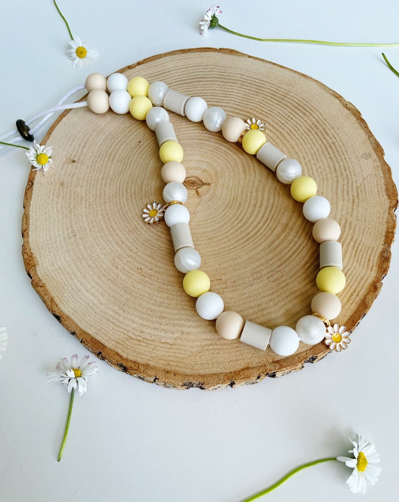 May include: A beaded lanyard featuring a sequence of round and cylindrical beads in shades of white, cream, beige, and yellow. The accessory includes small metal daisy charms with white petals and yellow centers, strung on a white cord.