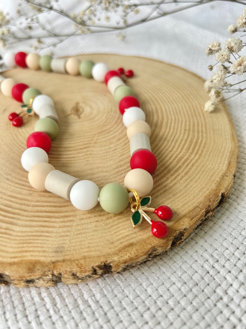 May include: A beaded necklace featuring a repeating pattern of round and cylindrical beads in shades of red, white, beige, and sage green. The necklace is accented with small gold-toned metal charms shaped like cherries with green enamel leaves.