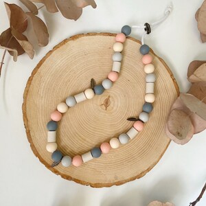 May include: A necklace featuring a repeating pattern of round and cylindrical beads in shades of slate grey, soft pink, and cream. The strand includes small metallic leaf-shaped charms and is secured with a white cord and a dark circular clasp.