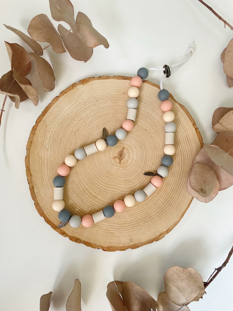 May include: A necklace featuring a repeating pattern of round and cylindrical beads in shades of slate grey, soft pink, and cream. The strand includes small metallic leaf-shaped charms and is secured with a white cord and a dark circular clasp.