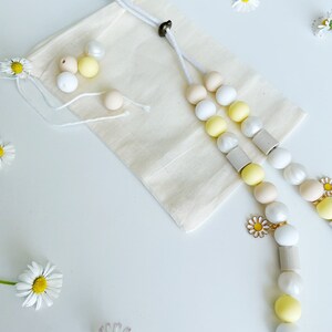 May include: A beige cotton drawstring pouch accompanied by a long beaded lanyard featuring white, yellow, and beige spherical beads, cylindrical wooden spacers, and a small metal daisy charm. Several loose beads and fresh white daisy flowers are present.