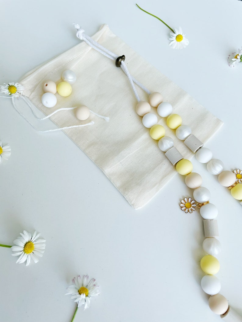 May include: A beige cotton drawstring pouch accompanied by a long beaded lanyard featuring white, yellow, and beige spherical beads, cylindrical wooden spacers, and a small metal daisy charm. Several loose beads and fresh white daisy flowers are present.