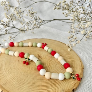 May include: A beaded necklace featuring a repeating pattern of round and cylindrical beads in shades of red, white, cream, and sage green. The necklace includes two gold-toned metal charms, each shaped like a pair of cherries with red enamel.