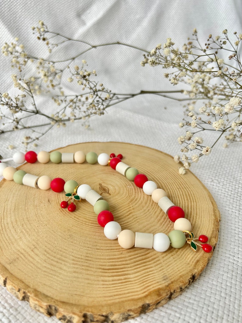 May include: A beaded necklace featuring a repeating pattern of round and cylindrical beads in shades of red, white, cream, and sage green. The necklace includes two gold-toned metal charms, each shaped like a pair of cherries with red enamel.