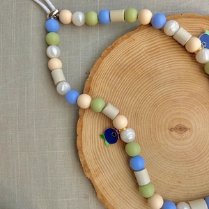 May include: A phone charm strap featuring a sequence of round beads in shades of light blue, sage green, cream, and white, interspersed with cylindrical wooden beads. The accessory includes a small, dark blue blueberry-shaped metal charm.