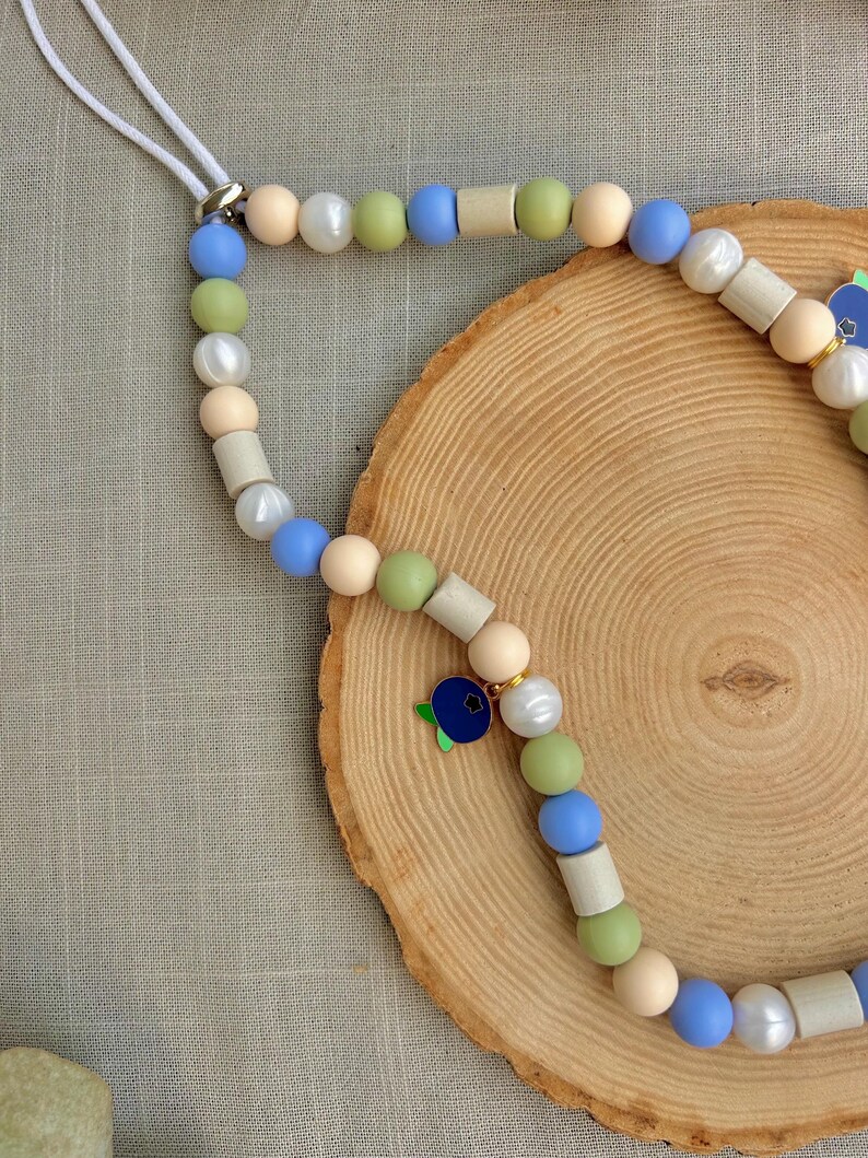 May include: A phone charm strap featuring a sequence of round beads in shades of light blue, sage green, cream, and white, interspersed with cylindrical wooden beads. The accessory includes a small, dark blue blueberry-shaped metal charm.