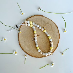 May include: A beaded lanyard featuring a mix of round and cylindrical beads in shades of white, yellow, and beige. The lanyard includes small daisy-shaped charms and is secured with a white cord and a dark brown adjustable toggle closure.