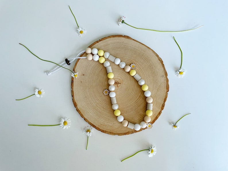 May include: A beaded lanyard featuring a mix of round and cylindrical beads in shades of white, yellow, and beige. The lanyard includes small daisy-shaped charms and is secured with a white cord and a dark brown adjustable toggle closure.
