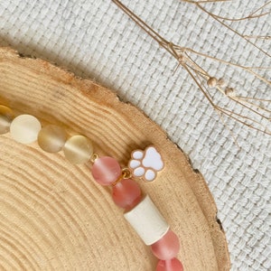 May include: A beaded bracelet featuring matte off-white cylindrical beads, translucent beige round beads, and translucent pink round beads. A small gold-toned charm shaped like a white paw print is attached to the bracelet between the beads.