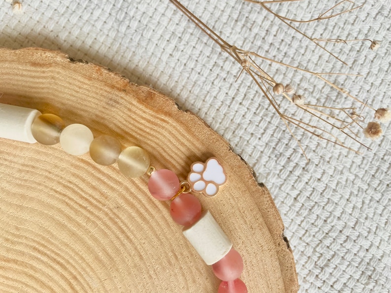 May include: A beaded bracelet featuring matte off-white cylindrical beads, translucent beige round beads, and translucent pink round beads. A small gold-toned charm shaped like a white paw print is attached to the bracelet between the beads.