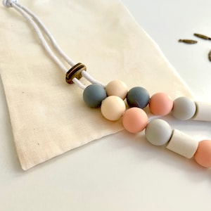 May include: A necklace featuring a series of smooth, matte-finish silicone beads in shades of slate grey, cream, and dusty rose, strung on a white cord with a bronze-toned adjustable slider bead, accompanied by a small, plain beige fabric pouch.
