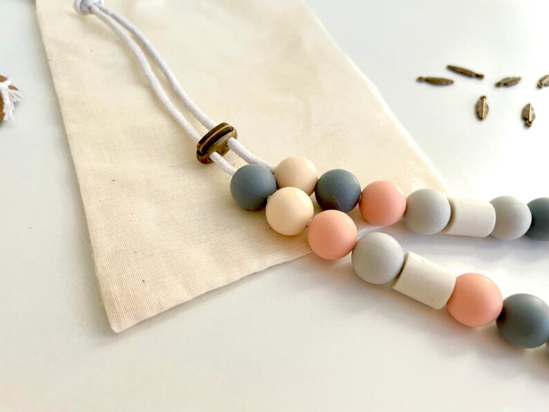 May include: A necklace featuring a series of smooth, matte-finish silicone beads in shades of slate grey, cream, and dusty rose, strung on a white cord with a bronze-toned adjustable slider bead, accompanied by a small, plain beige fabric pouch.