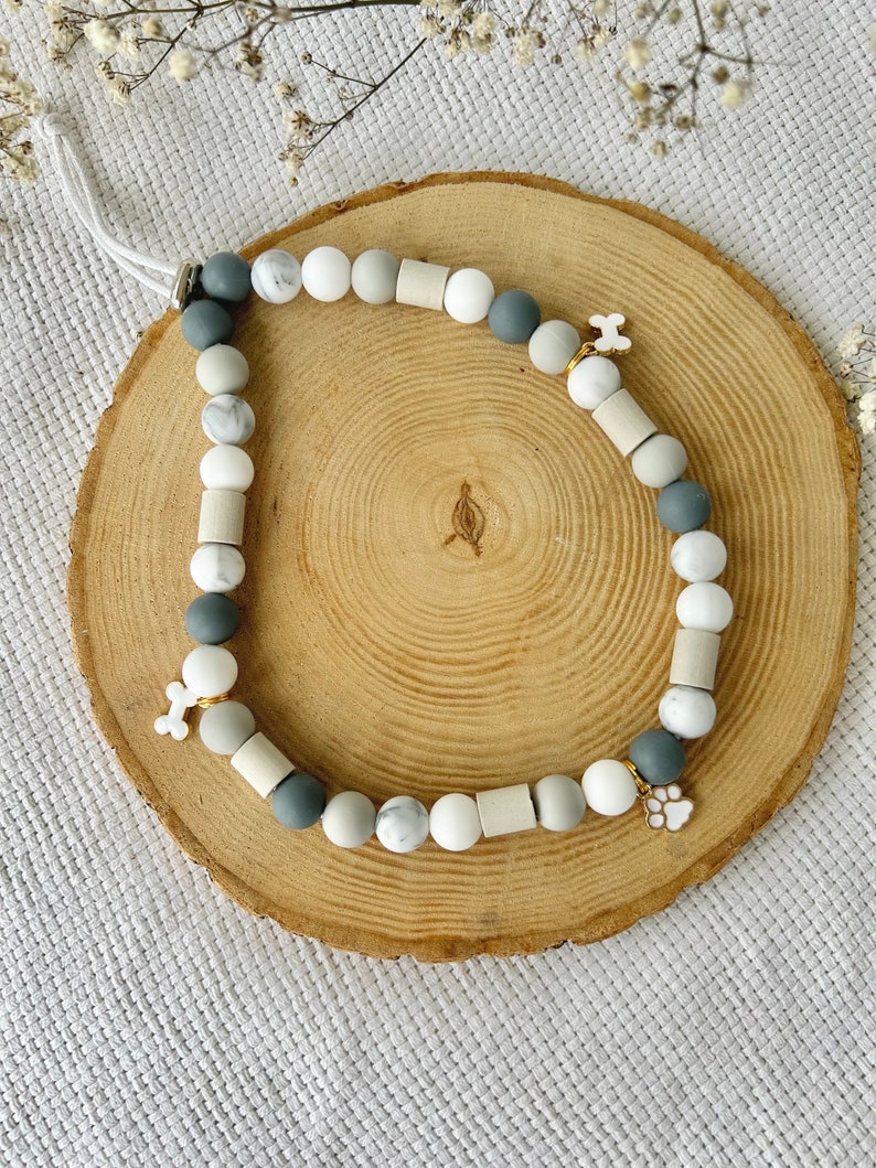 May include: A beaded accessory featuring a mix of round and cylindrical beads in shades of white, grey, and marble patterns. The strand includes two white bone-shaped charms and one white paw print charm, all strung on a white cord with a metal clasp.