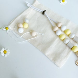 May include: A beige cotton drawstring pouch accompanied by a beaded accessory featuring round white, yellow, and tan beads, along with square grey beads. Several loose beads in matching colors are scattered near the pouch, which is decorated with white daisy flowers.